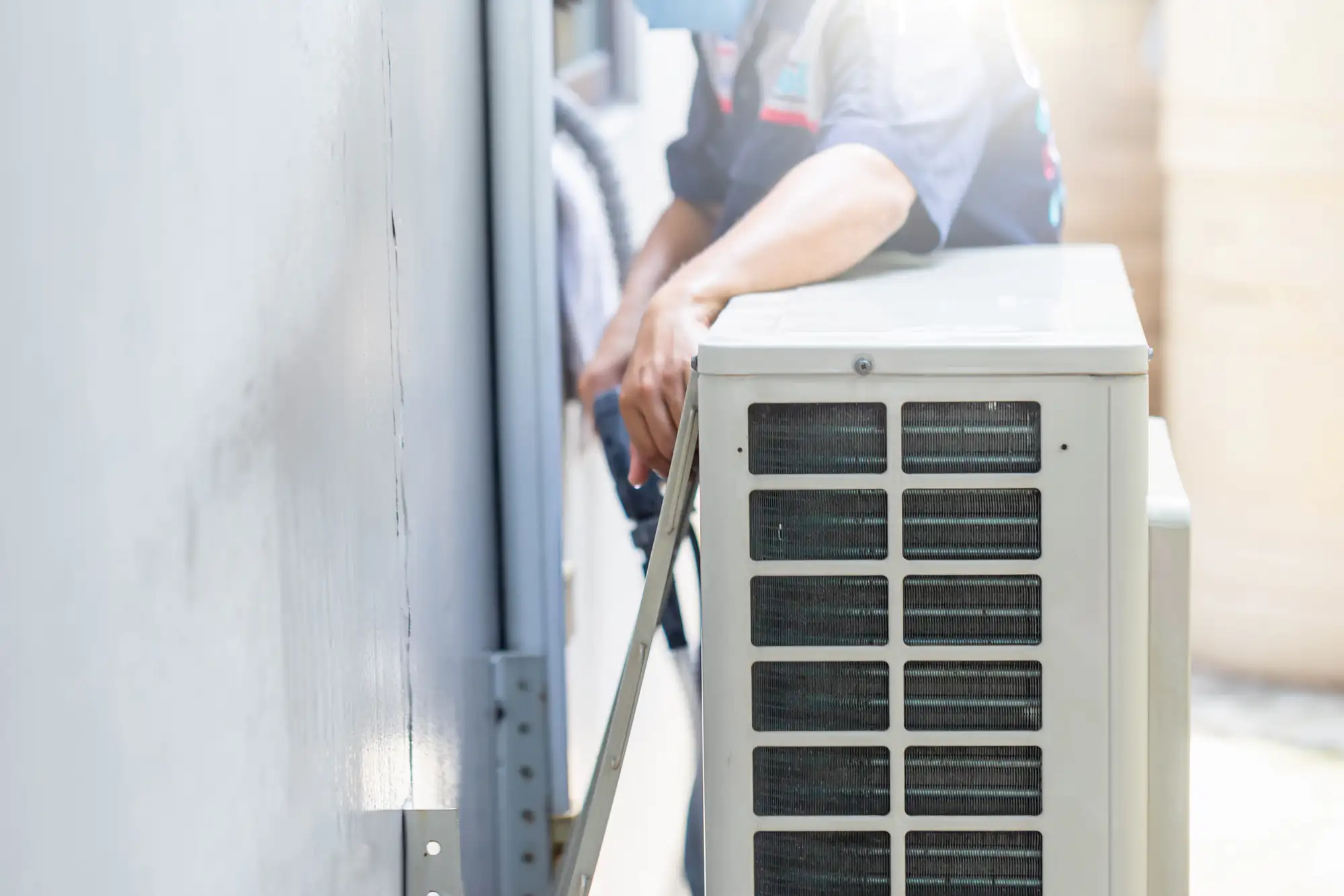 A technician in TX installs or repairs an outdoor air conditioning unit mounted against a wall, tools in hand and the unit's vented side visible&mdash;ideal for ac replacement Bexar County residents can rely on.