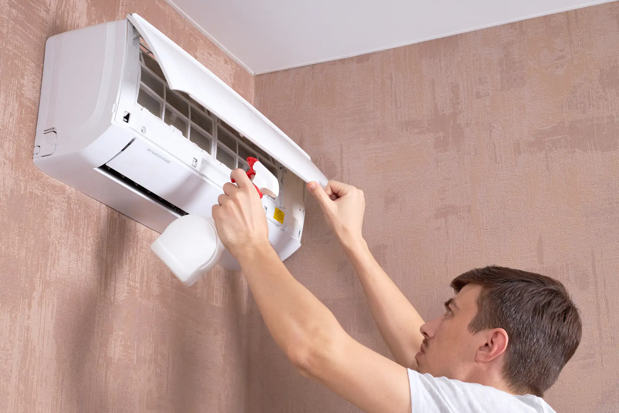 A man in TX inspects and cleans an open wall-mounted air conditioner with a spray bottle, standing close to a textured beige wall&mdash;essential maintenance before considering ac replacement Bexar County homeowners may need.