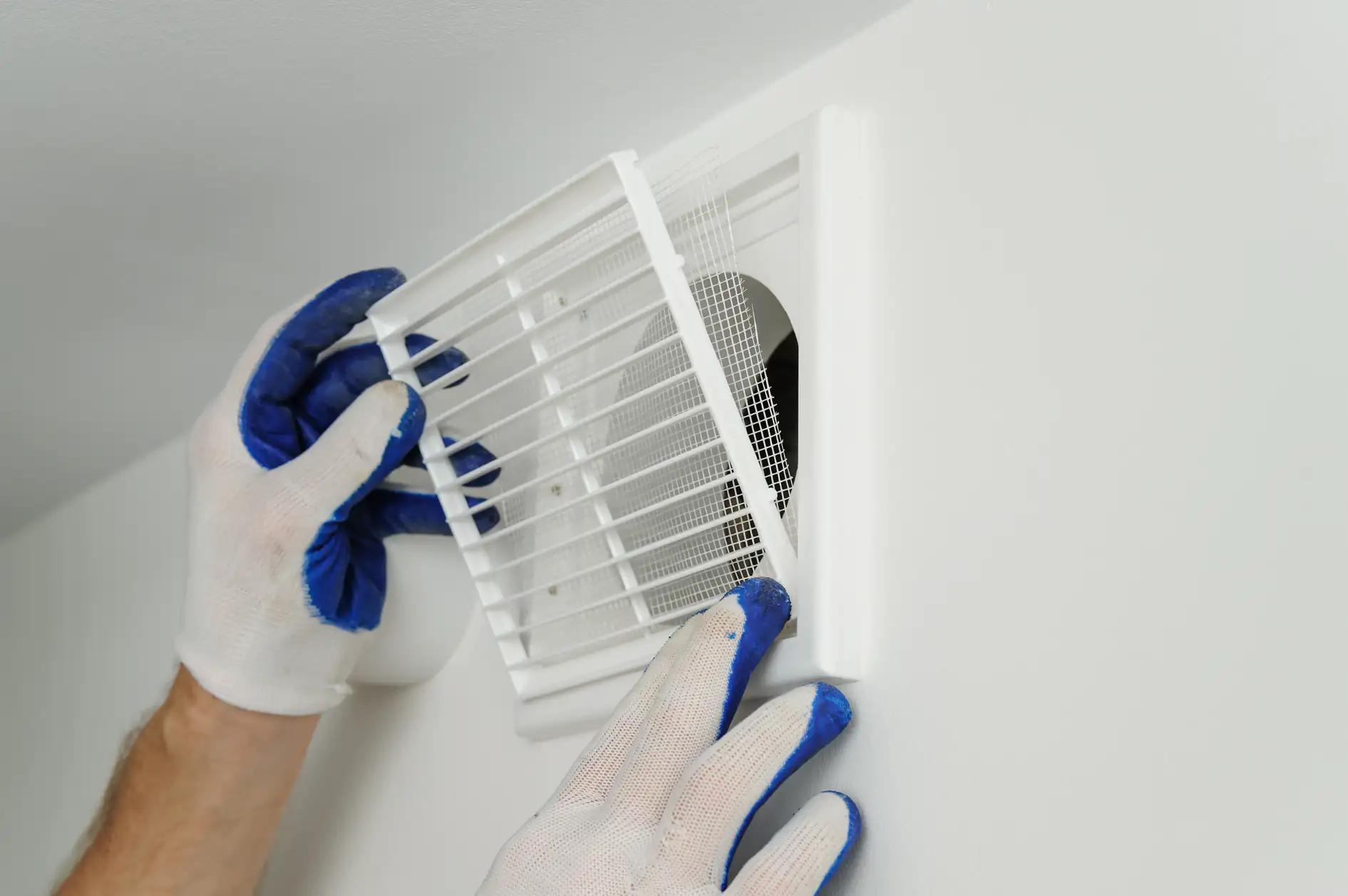 A person wearing white gloves with blue fingertips is installing or removing a white air vent cover from a wall in TX, likely as part of an ac replacement Bexar County project. The vent cover is held at an angle, revealing the duct opening behind it.