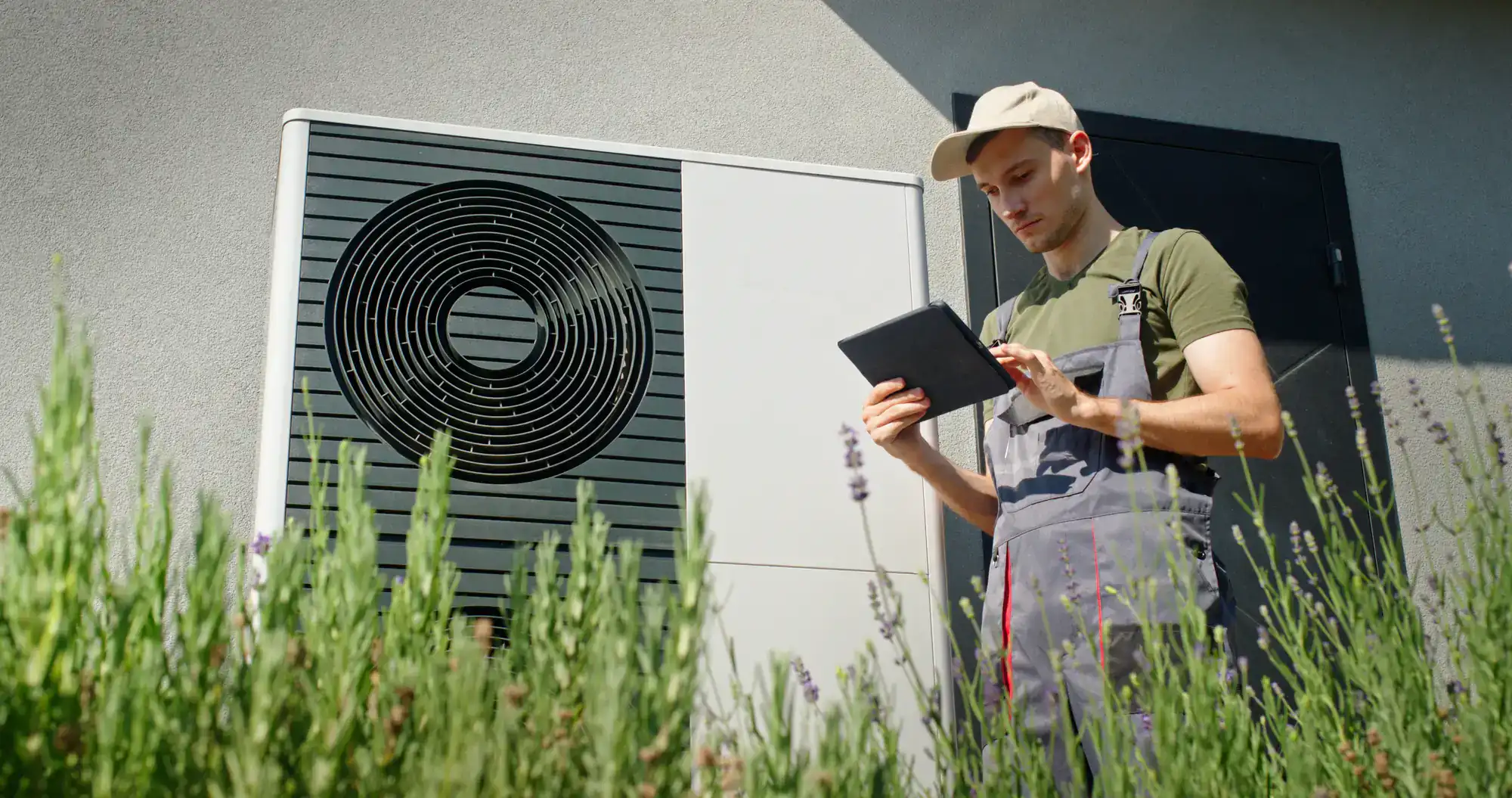 A technician in overalls and a cap stands outdoors in front of a heat pump unit in Bexar County, TX, using a digital tablet. Tall green plants are in the foreground, and a building wall is in the background&mdash;ideal for ac replacement projects.