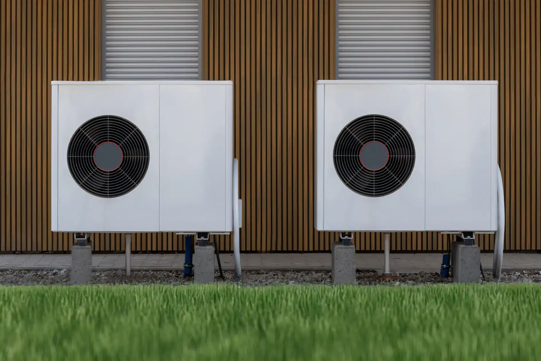 Two white outdoor heat pump units with large black fans stand side by side on concrete blocks in front of a modern building with vertical wooden slats and closed metal shutters in TX&mdash;ideal for ac replacement Bexar County residents can rely on. Green grass is visible in the foreground.