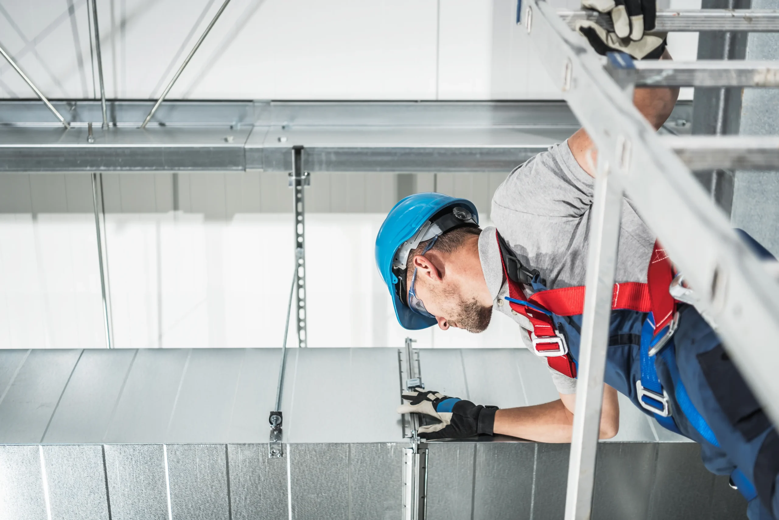 A construction worker wearing a blue hard hat, gloves, and a safety harness installs or inspects metal ductwork on a ceiling&mdash;possibly for an AC replacement Bexar County, TX project&mdash;while standing on a ladder inside an industrial or commercial building.