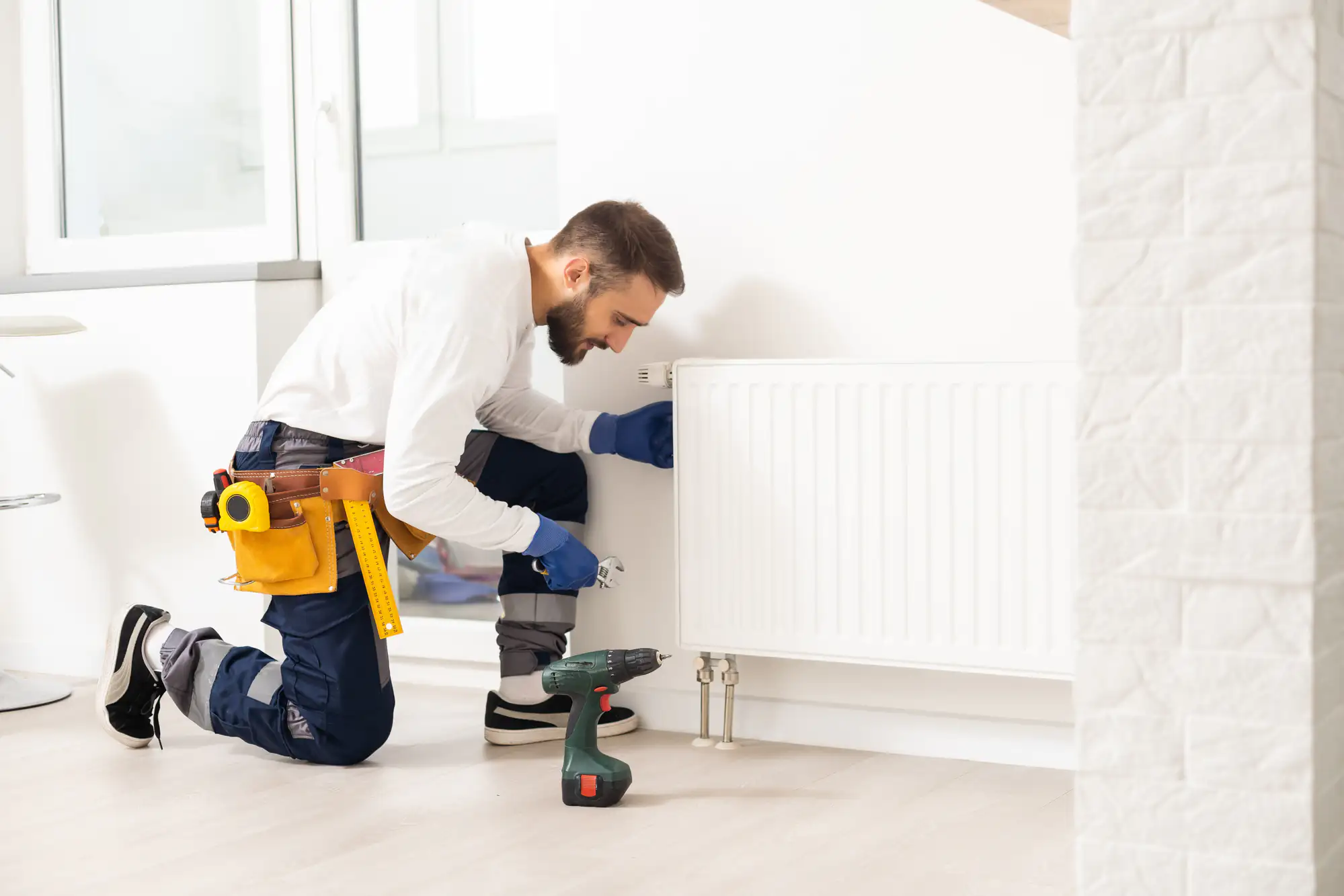 A man in work clothes and a tool belt kneels on the floor, repairing or installing a white radiator in a bright room. Nearby, a power drill and various tools suggest a TX ac replacement Bexar County project is underway.