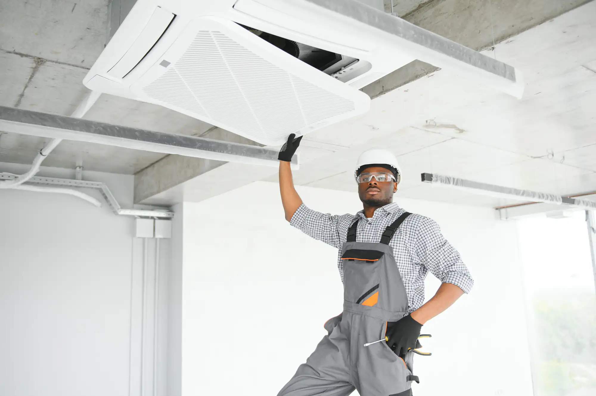 A technician in overalls, safety glasses, gloves, and a hard hat inspects or repairs a ceiling-mounted air conditioning unit in a modern, unfinished room&mdash;perfect for TX summers or ac replacement in Bexar County.