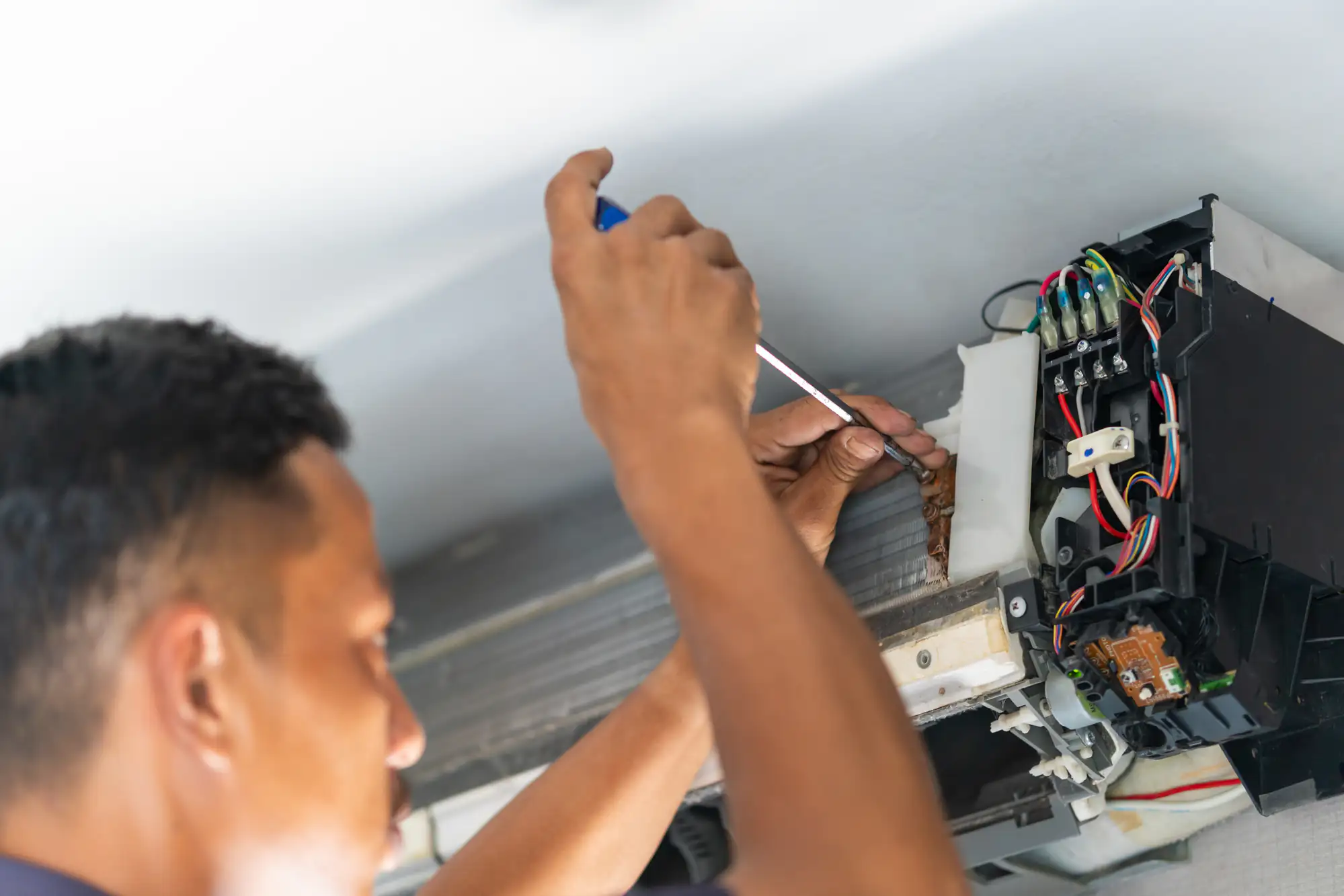 A person in TX uses a screwdriver to repair or maintain the internal components of a wall-mounted air conditioning unit, with exposed wires and parts visible&mdash;common during ac replacement Bexar County residents may need.