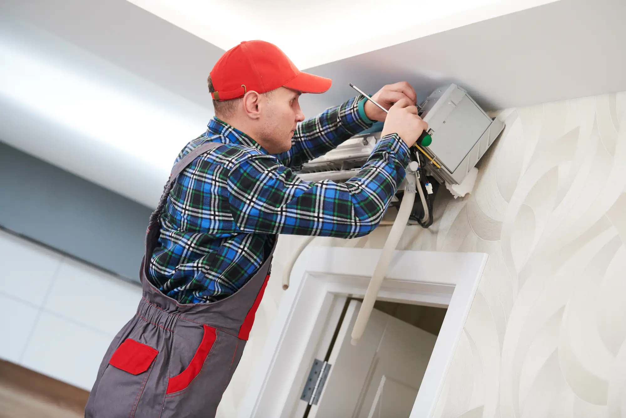 A technician wearing a red cap, plaid shirt, and overalls repairs an air conditioning unit mounted high on a wall inside a TX home, using tools and a screwdriver&mdash;ideal for AC replacement Bexar County residents may need.