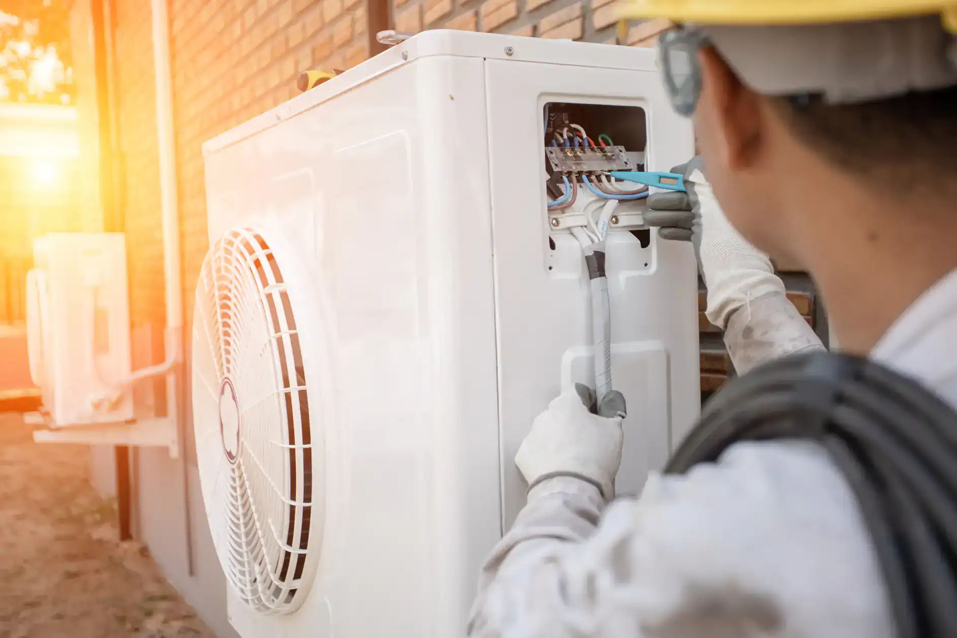 A technician wearing safety gear uses tools to repair or install wiring on an outdoor air conditioning unit next to a brick wall in bright sunlight, providing expert heating services in Bexar County, TX.