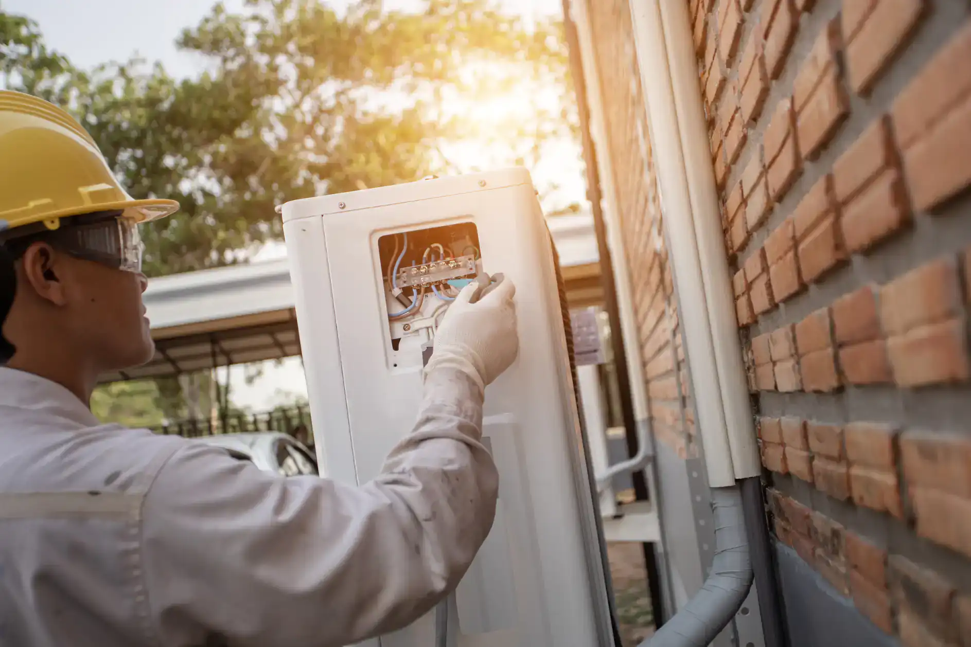A worker wearing a yellow hard hat and white gloves repairs or installs an electrical unit on a building&rsquo;s brick wall in Bexar County, TX, with sunlight shining in the background&mdash;possibly part of an ac replacement project.