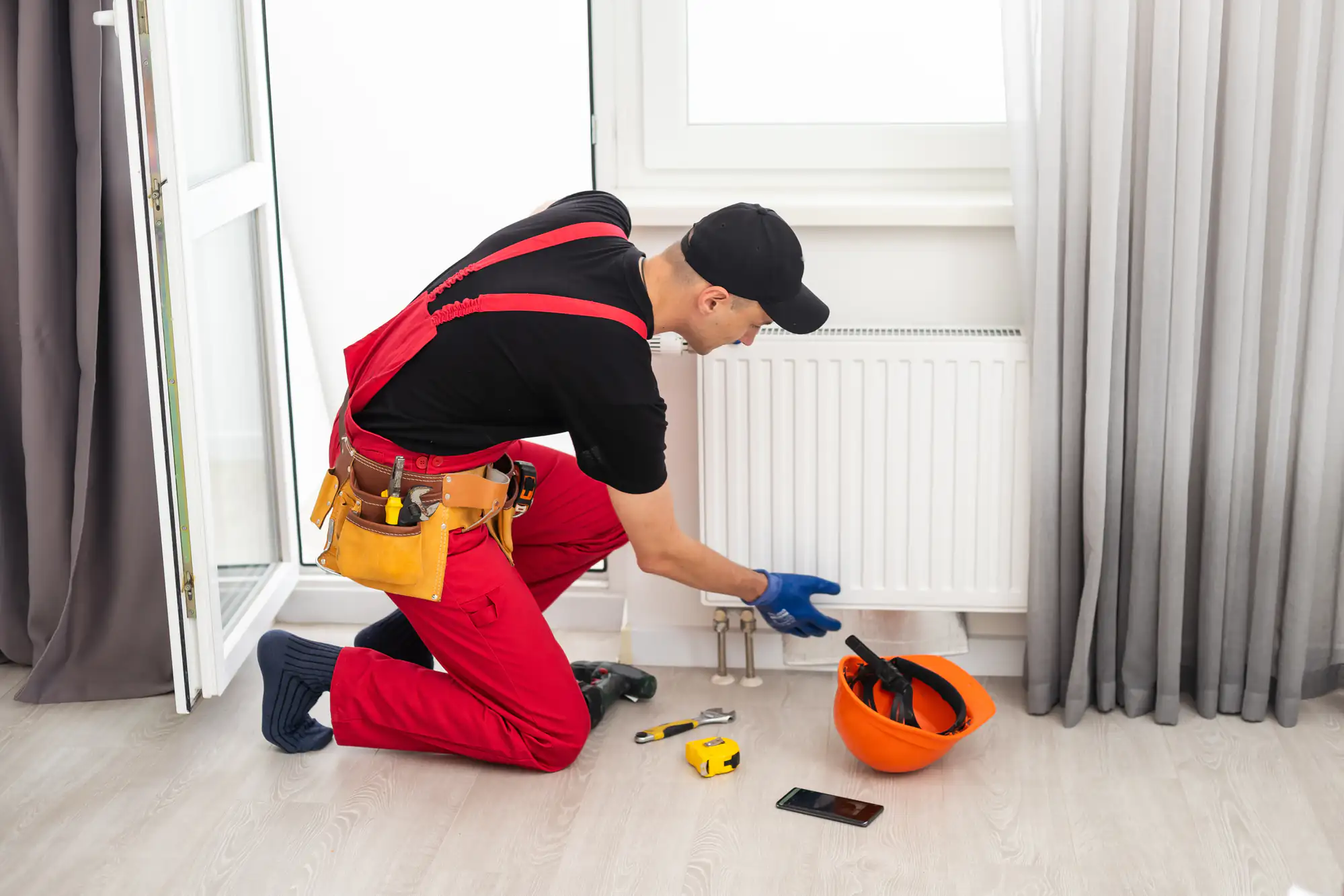A plumber in red overalls and a black cap kneels on the floor, inspecting a white radiator. Tools, a phone, and an orange hard hat with more tools are on the floor beside him, ready for any TX repairs or ac replacement Bexar County calls.