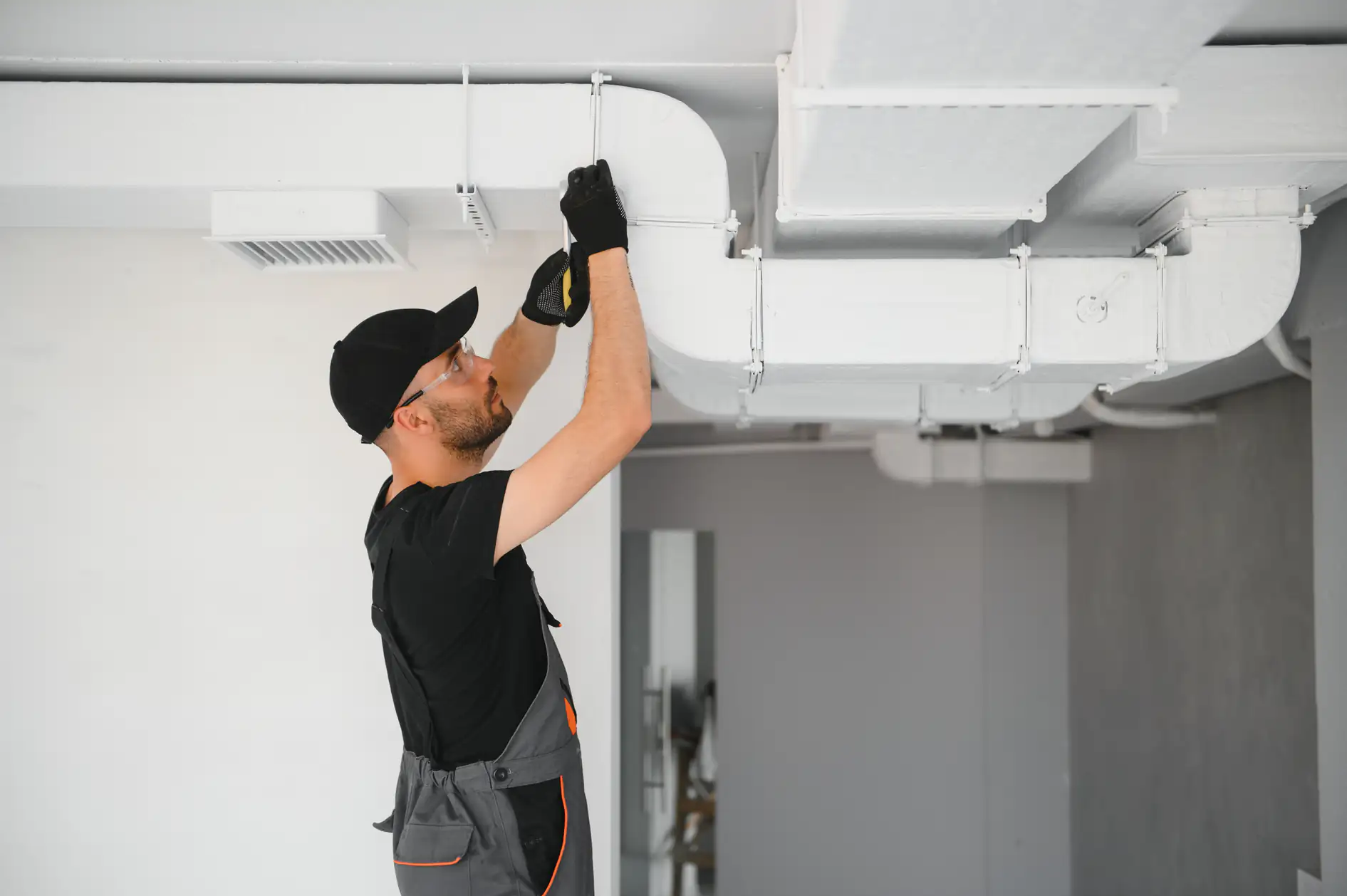 A worker in a black cap and gloves uses a screwdriver to fix or install an overhead ventilation duct, part of an AC replacement in a modern industrial interior in Bexar County, TX.